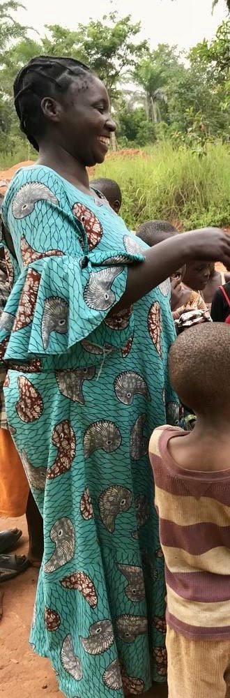 A Tanzanian woman in a blue kitenge dress smiles as she hands out supplies; the children in front of her are seen from behind